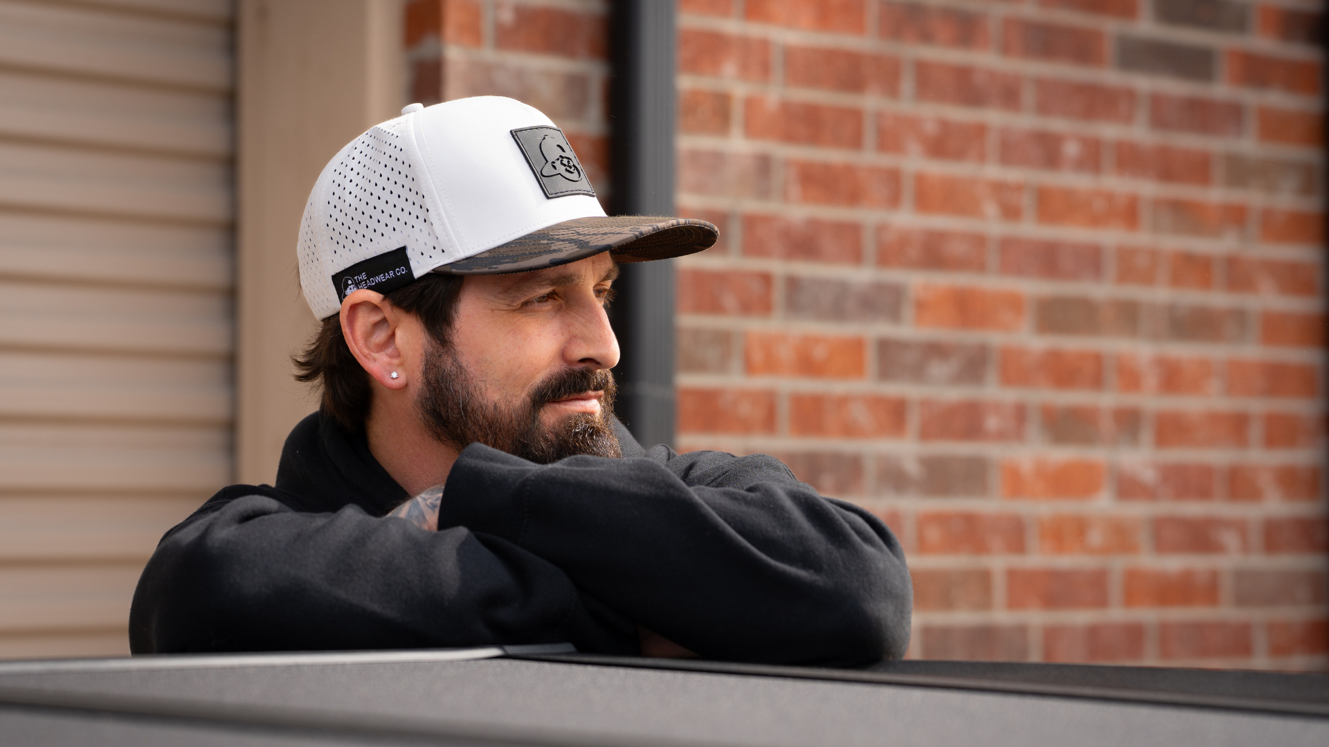 Man wearing a white cap with a logo, leaning against a vehicle with a brick wall background